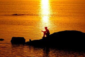 negro silueta de un ciclista a puesta de sol con un reflexión en el agua con ondas descanso en rocas sentado cerca agua. foto