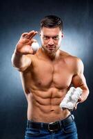 Handsome young sportsman is holding a packaging with eggs, healthy food protein, gray background. showing one egg to camera. Naked torso, healthy life concept. photo