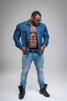 Handsome African guy with naked torso wearing jeans shirt. Athletic man in denim sanding on white background. Bodybuilder male model wearing glasses. photo