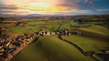 Aerial view of a scenic countryside at sunset with lush green fields, a small village, and a winding road leading towards distant hills. photo