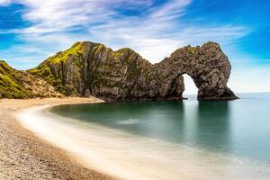 Scenic view of a natural arch on a coastline with a sandy beach and clear blue sky at Durdle Door photo