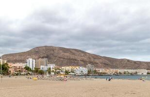 Scenic view of a sandy beach with a mountain backdrop under a cloudy sky, with buildings along the coastline in Los Cristianos, Tenerife. photo