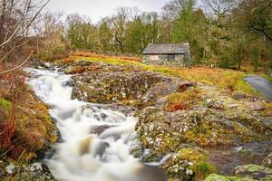 Rustic stone cottage by a flowing river in a serene woodland setting, showcasing vibrant autumn foliage and a tranquil atmosphere in Lake District. photo