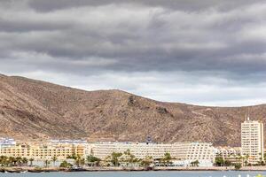 Coastal cityscape with hotels against mountains under cloudy skies in Los Cristianos, Tenerife. photo