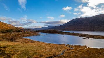Serene landscape with a calm lake, rolling hills, and a clear blue sky with clouds, reflecting the beauty of untouched nature in Scotland. photo
