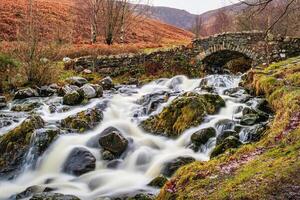 Rustic stone bridge over a flowing stream with mossy rocks in a lush, green landscape in Lake District. photo