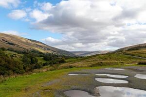 Scenic view of a lush valley with rolling hills under a cloudy sky, featuring a road with puddles after rain. photo