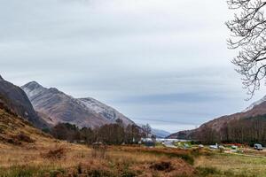 Serene valley landscape with mountains under a cloudy sky, bare tree branches on the side, and a glimpse of a water body in the distance. photo