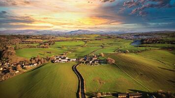 aéreo ver de un pintoresco pueblo en medio de verde campos durante puesta de sol con un vibrante cielo y un serpenteante río. foto