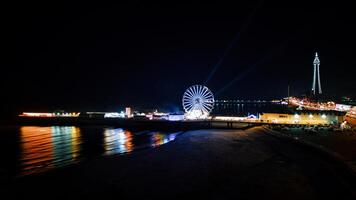 Coastal night scene with illuminated Ferris wheel and lighthouse, vibrant lights reflecting on water, and a dark beach foreground in Backpool, England. photo