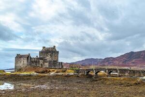 Medieval castle with stone bridge against a dramatic sky, surrounded by rugged landscape and autumn colors in Scotland. photo