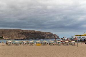 Cloudy day at the beach with empty sun loungers and umbrellas, with cliffs in the background in Los Cristianos, Tenerife. photo