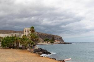 Coastal landscape with beach, palm trees, and hotels against a backdrop of cliffs under a cloudy sky in Los Cristianos, Tenerife. photo