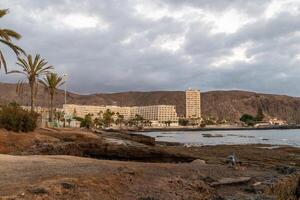 Coastal landscape with hotels, palm trees, and mountains under a cloudy sky in Los Cristianos, Tenerife. photo