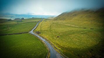 Aerial view of a winding road through lush green hills with mist and a glimpse of the ocean in the distance. photo