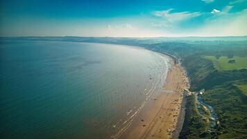 Aerial view of a sweeping coastline with a sandy beach, calm sea, and greenery under a blue sky with scattered clouds in Filey, England. photo