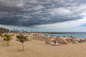 Cloudy sky over a busy beach with people sunbathing and a pier in the distance in Los Cristianos, Tenerife. photo