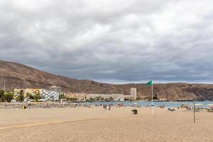 Sandy beach with tourists, green flag, calm sea, cloudy sky, and coastal buildings in the background in Los Cristianos, Tenerife. photo