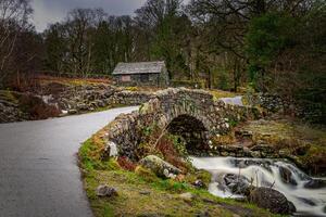 Stone bridge over a stream with a cottage in the background, surrounded by a lush landscape in Lake District. photo