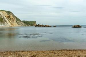 Tranquil beach scene with calm waters, sandy shore, and rocky cliffs under a soft cloudy sky at Durdle Door, England. photo