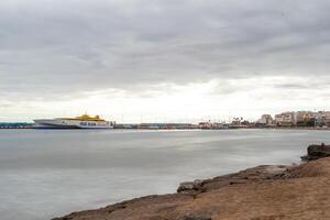 Serene seascape with a ferry boat on the horizon under a cloudy sky, coastal cityscape in the background in Los Cristianos, Tenerife. photo