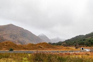 Serene Scottish Highlands landscape with misty mountains, golden grass, and a small cottage beside a road. photo