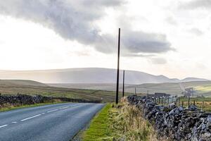 Empty countryside road with a telephone pole, stone walls, and rolling hills under a cloudy sky in Yorkshire Dales. photo