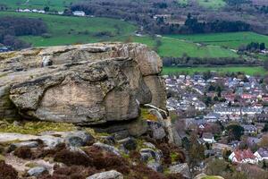 Scenic view of a large rock formation overlooking a lush valley with a small town and green fields in the background in Ilkley, Yorkshire photo