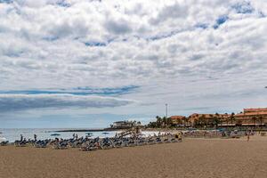 Serene beach scene with cloudy sky, sun loungers, and umbrellas on sandy shore, with calm ocean and coastal buildings in the background in Los Cristianos, Tenerife. photo