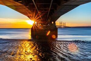 Sunset view under a pier with sunburst effect, reflecting on wet sand and calm sea. photo