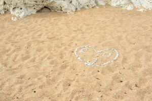 corazón forma hecho de conchas marinas en arenoso playa con rocas en antecedentes. foto