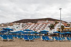 Cloudy day at a beach resort with rows of blue sun loungers and white buildings on a hill in the background in Los Cristianos, Tenerife. photo