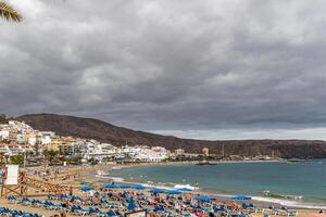 Cloudy day at a beach resort with rows of blue sun loungers and umbrellas, with a town on the hillside in the background in Los Cristianos, Tenerife. photo