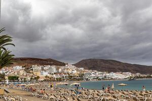 nublado día a un playa recurso con turistas relajante en Dom tumbonas, palma árbol en primer plano, y costero pueblo fondo en los cristianos, tenerife foto