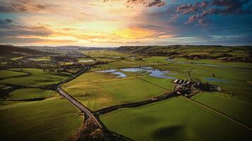 Aerial view of a scenic countryside at sunset with vibrant skies, lush green fields, and a winding road leading to a farmhouse. photo