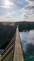 Aerial view of a narrow bridge over a tranquil river, surrounded by lush greenery under a partly cloudy sky. photo