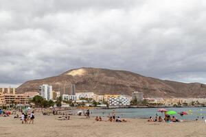 Scenic view of a beach with people relaxing, cityscape, and a large mountain under cloudy skies in Los Cristianos, Tenerife. photo