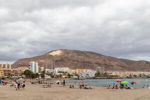 Scenic view of a beach with people relaxing, cityscape, and a large mountain under cloudy skies in Los Cristianos, Tenerife. photo