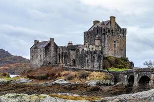 Majestic medieval castle with stone bridge over a river, surrounded by wild nature under a cloudy sky in Scotland. photo