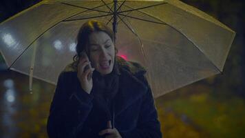 enojado joven mujer hablando en inteligente teléfono en lluvioso ciudad a noche discutiendo video