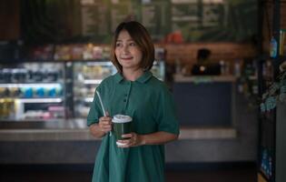 mujer en pie en el café participación un bebida con un papel Paja y café taza, sonriendo y mirando a el cámara, relajante en vacaciones, estilo de vida conceptos. foto