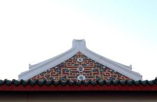 Ancient native Chinese style art in Thailand on isosceles triangle shape gable of church in temple white sky background. White cement rim and brown geometric pattern design on gable. photo