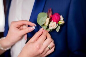 Bride's hands pinning a small boutonniere to a jacket for the groom. Floral traditional decoration for groom, an accessory on festive wedding suit. Close-up photo