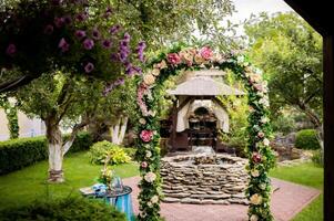 Beautiful arch decorated with colorful flowers on the background of a small fountain outdoors. Natural background in the park with floral archway, green grass and trees in a sunny day. photo