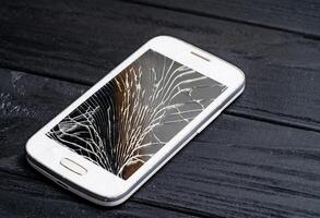 Modern cellphone with broken touchscreen isolated on black surface. Destroyed white mobile phone lying on the floor. Close-up photo