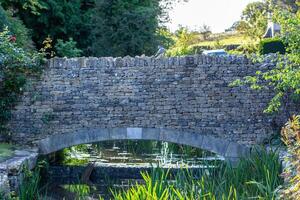 Old stone bridge over a tranquil stream with lush greenery in a serene countryside setting. photo