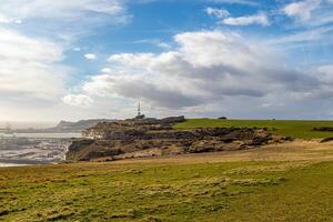 Coastal landscape with green fields under a cloudy sky, overlooking a rocky shoreline and a distant lighthouse. photo