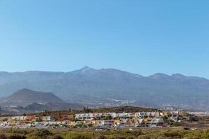 Scenic view of a tranquil town with white buildings nestled at the foot of a mountain range under a clear blue sky in Tenerife. photo