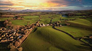 Aerial view of a quaint village amidst lush green fields with rolling hills in the background during sunset. photo