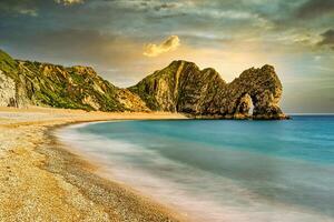 sereno playa con suave aguas, dorado arena, y escabroso acantilados debajo un puesta de sol cielo a durdle puerta, Inglaterra. foto